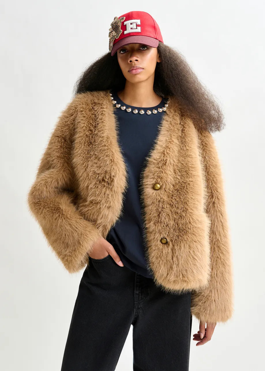 A person wearing the Essentiel Antwerp Ijacket Faux Fur Jacket in brown, a black shirt with a decorative collar, black jeans, and a red cap with an "E" emblem stands against a plain light background.