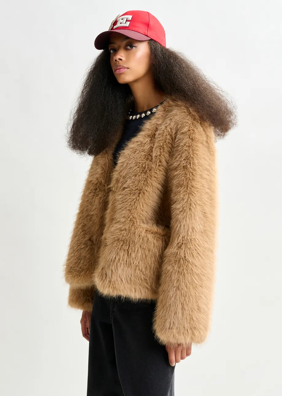 A woman wears the Essentiel Antwerp Ijacket Faux Fur Jacket in brown, paired with black pants and a red baseball cap, standing against a plain light background.