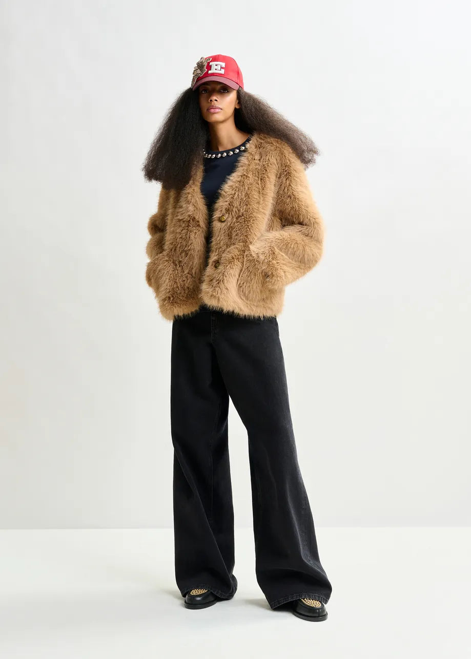 A woman wears the Essentiel Antwerp Ijacket Faux Fur Jacket in brown, styled with a black shirt, wide-leg black pants, and a red baseball cap, standing against a plain white background.
