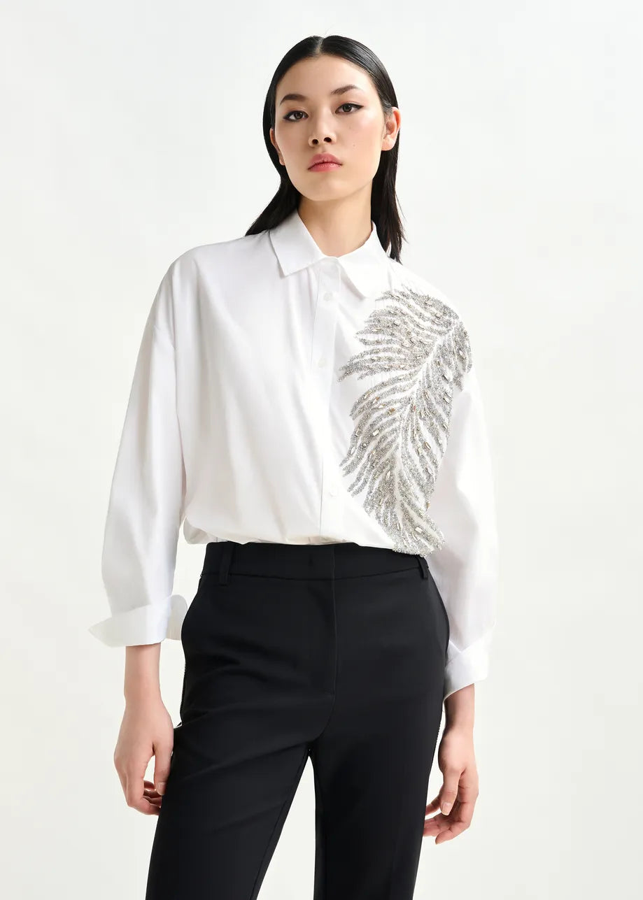 A person wears the Essentiel Antwerp Iliza Sequin Shirt in white, featuring a silver sequin feather design on one side, paired with black pants and standing against a plain background.