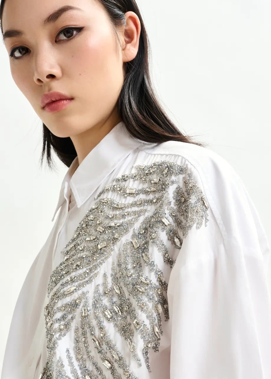 A person wearing the Iliza Sequin Shirt in white by Essentiel Antwerp, featuring oversized cotton and intricate silver sequin embroidery, poses against a plain light background.