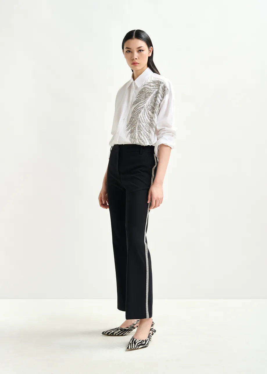 A woman wears the Essentiel Antwerp Iliza Sequin Shirt in white, paired with black side-stripe pants and zebra-print shoes, standing against a plain white background.