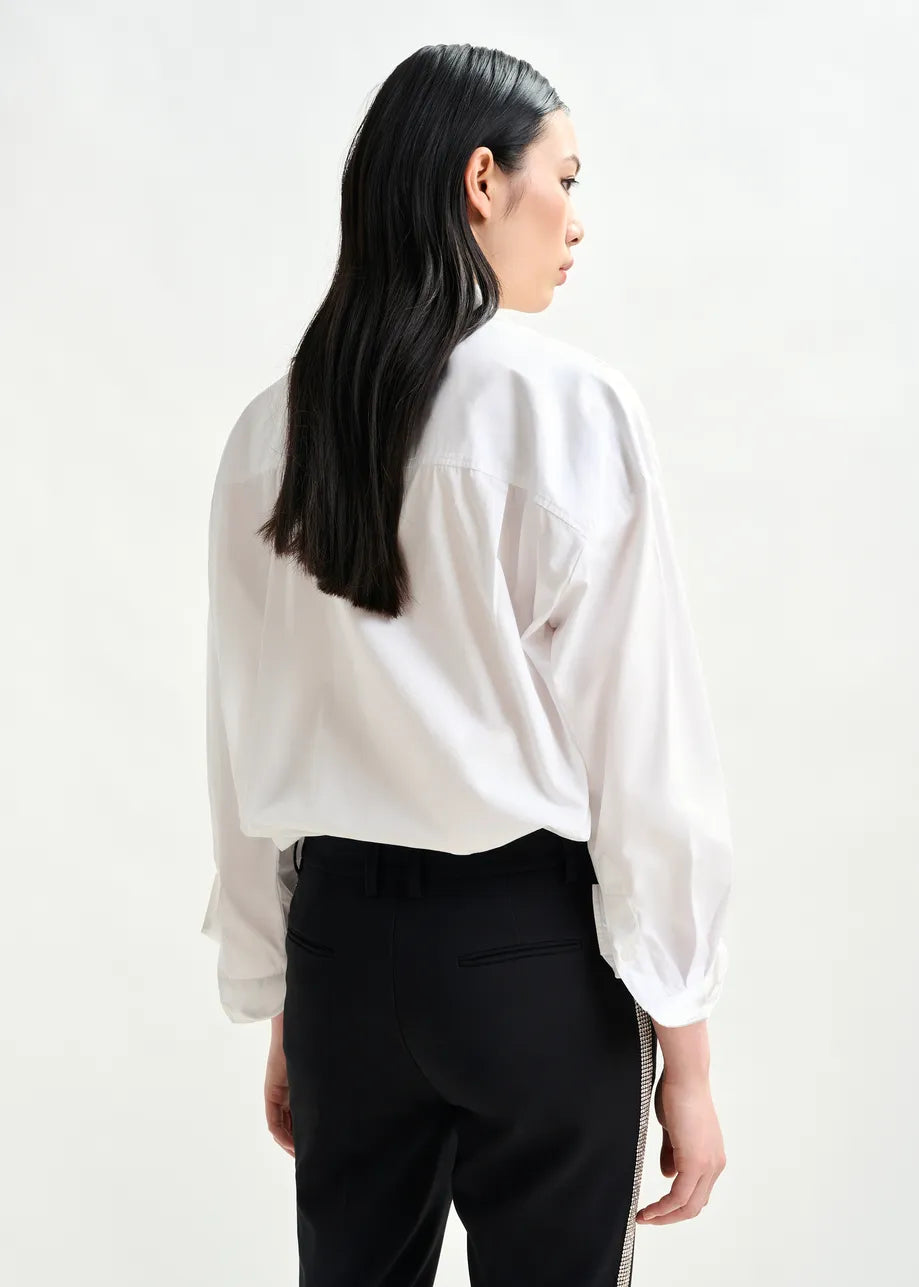 A person with long dark hair stands facing away, wearing the Iliza Sequin Shirt in white by Essentiel Antwerp and black pants against a plain light background.