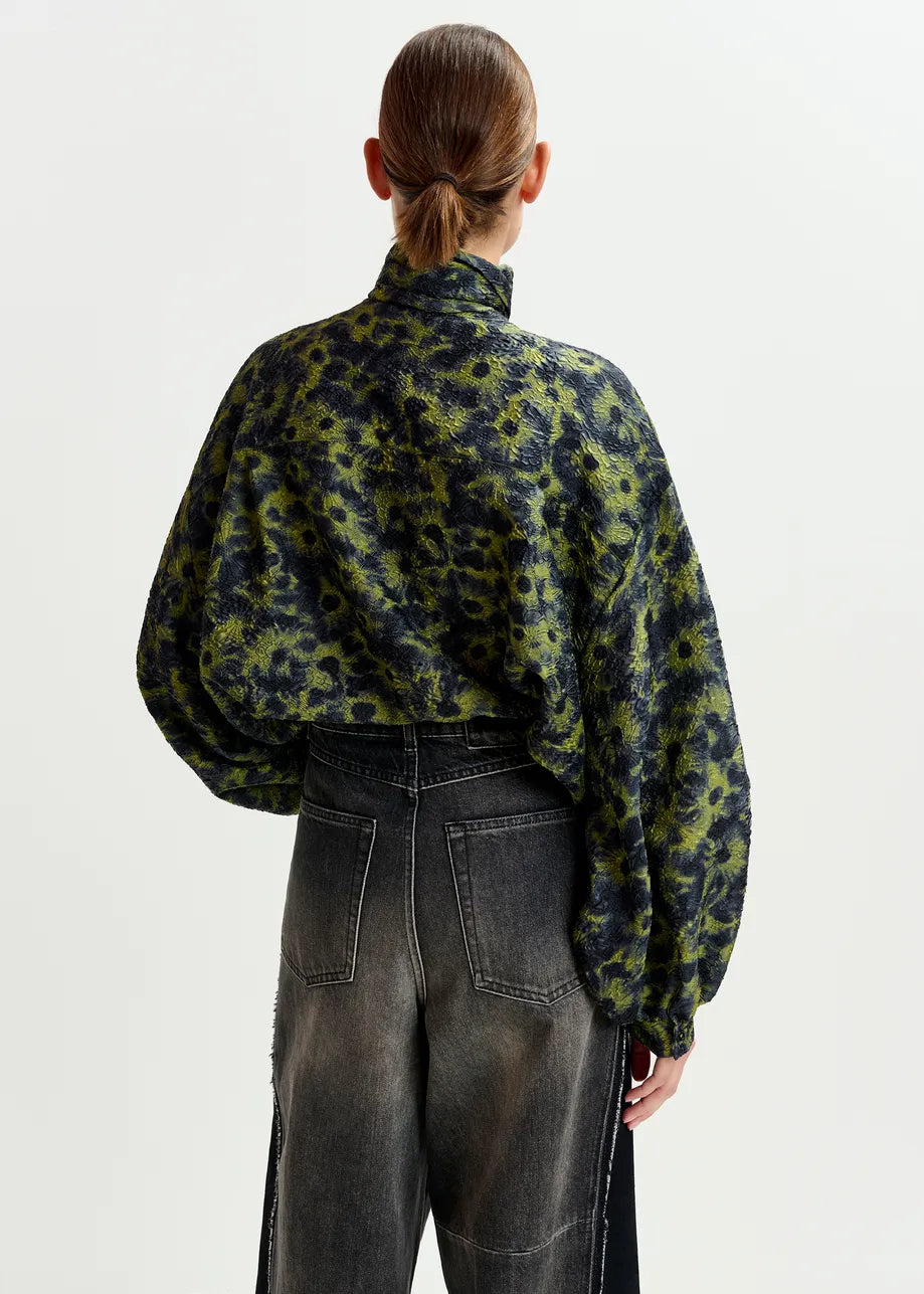 A person stands with their back to the camera, wearing the Illicium Floral Jacquard Shirt in khaki by Essentiel Antwerp, paired with high-waisted, faded black jeans.