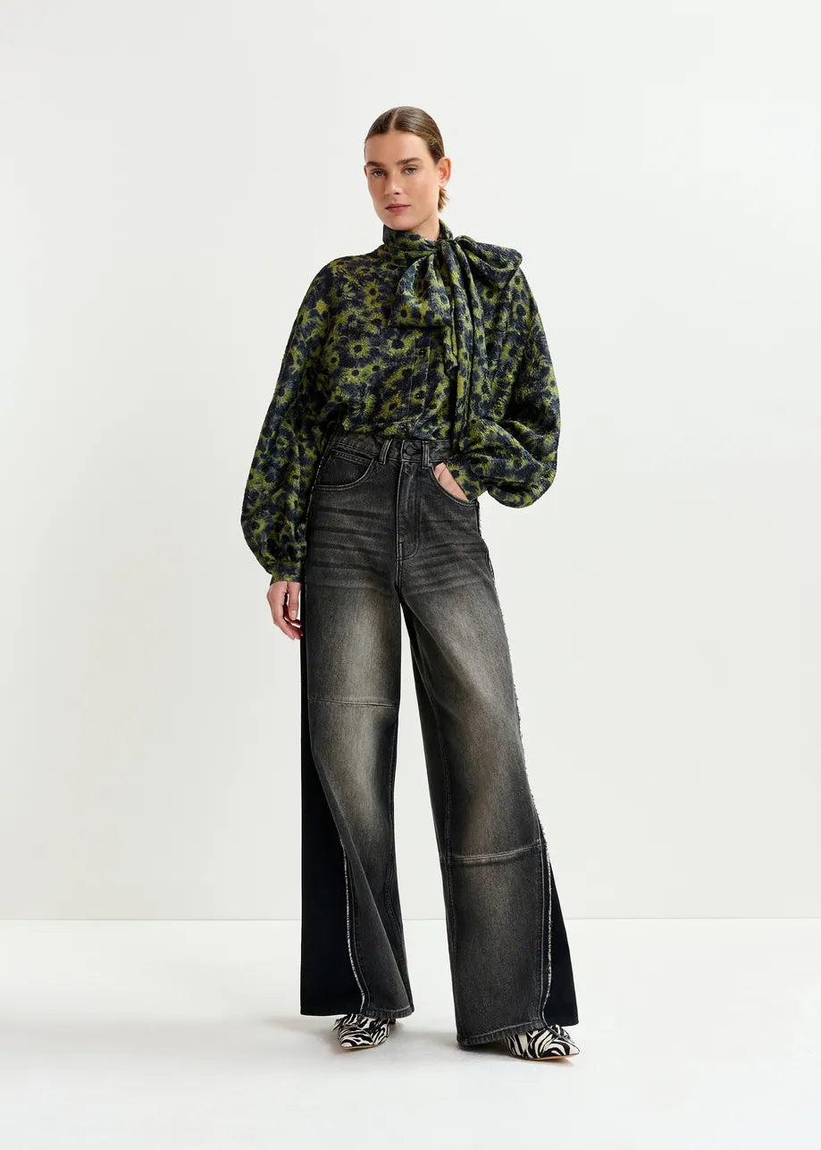 A person stands against a plain background wearing the Essentiel Antwerp Illicium Floral Jacquard Shirt in khaki, paired with high-waisted wide-leg faded black jeans and patterned shoes.