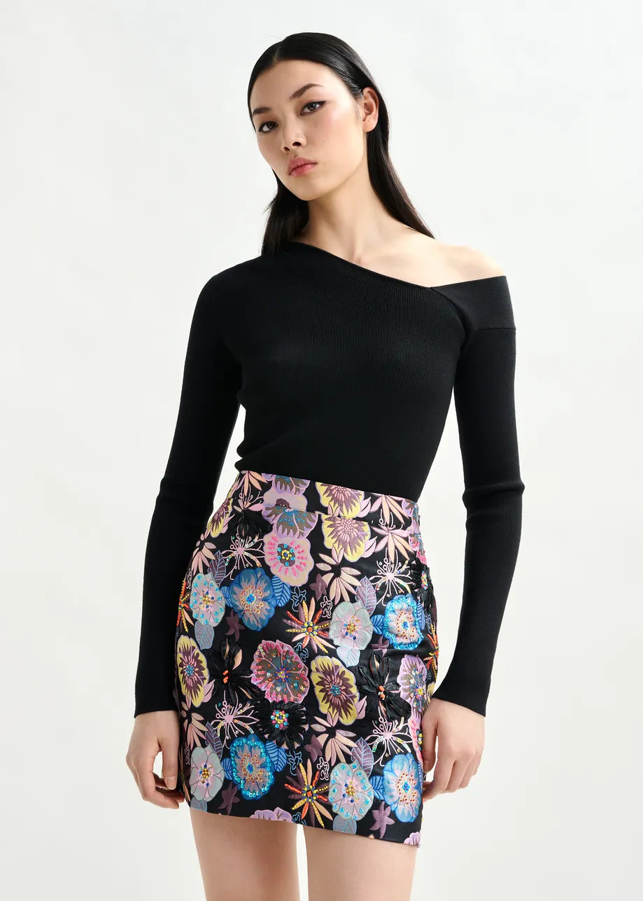 A woman wears the Essentiel Antwerp Imeric Asymmetrical Jumper in black with an off-the-shoulder neckline, paired with a high-waisted, floral-print mini skirt, standing against a plain light background.