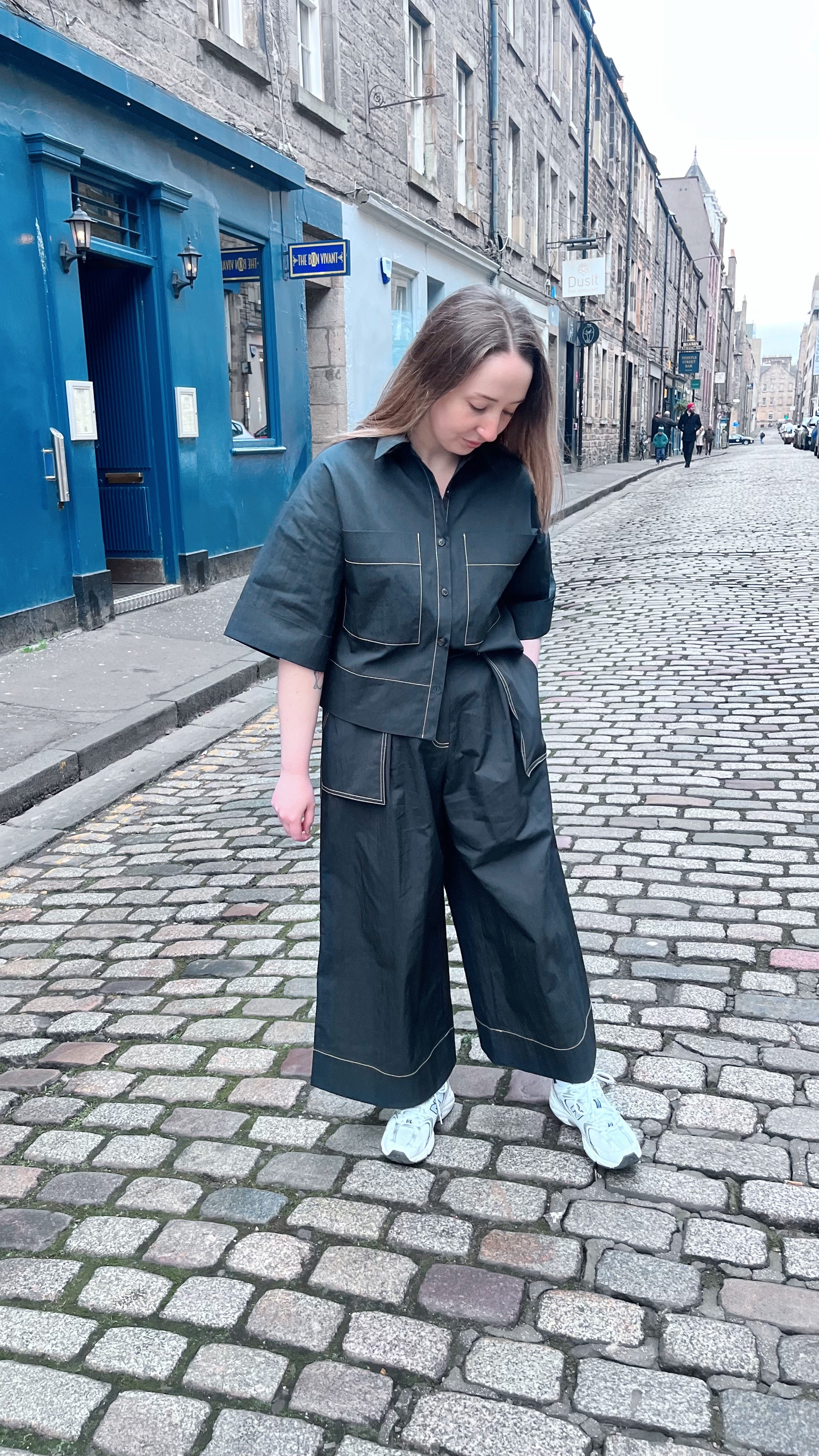 A person wearing Emin and Paul Wide Leg Trousers in black and white sneakers stands on a cobblestone street, looking down at their feet. Buildings line the background.