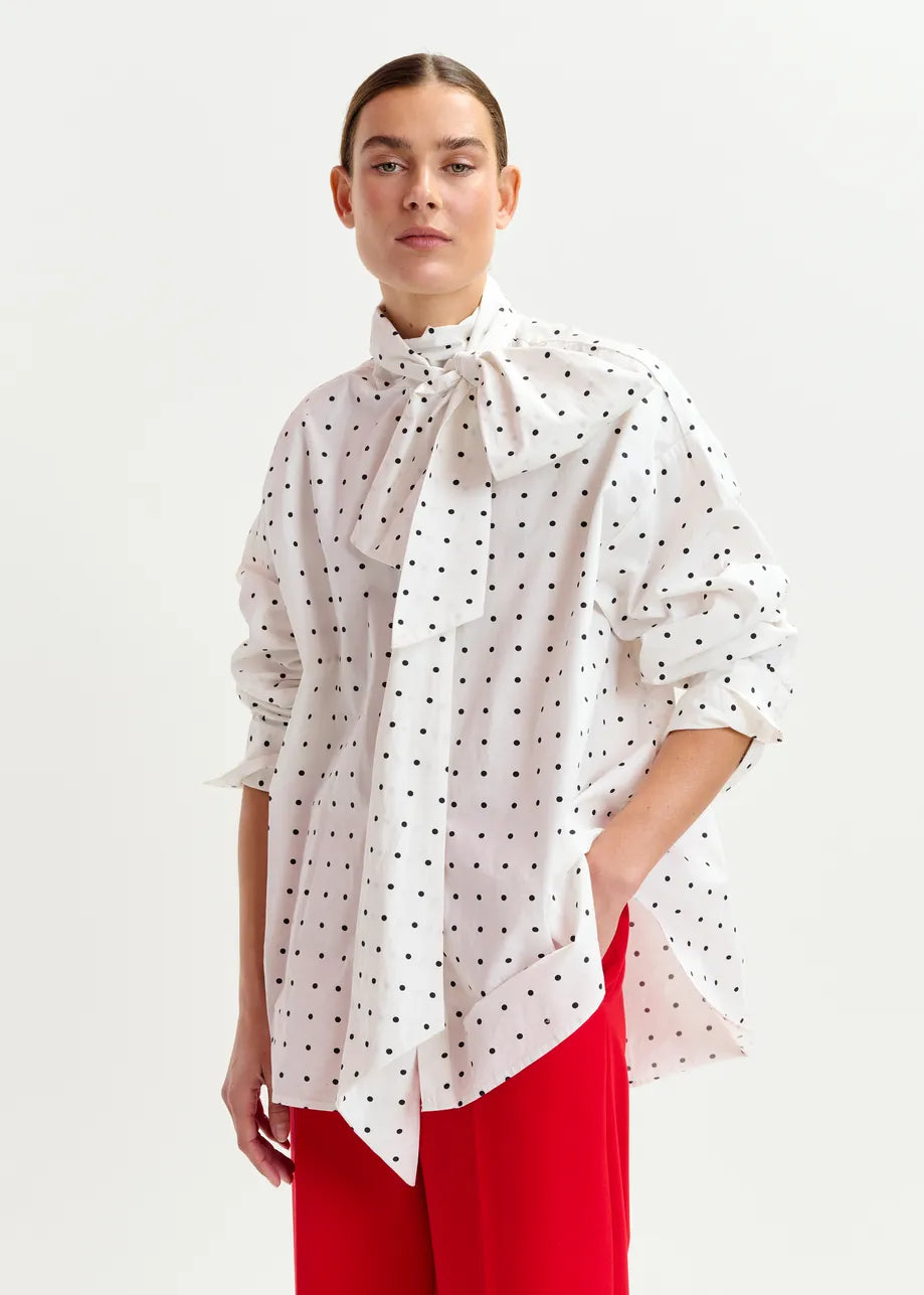 A person wears the Essentiel Antwerp Imoverit Polkadot Shirt in white and black, featuring a large bow at the neck, paired with red pants, and stands against a plain white background.