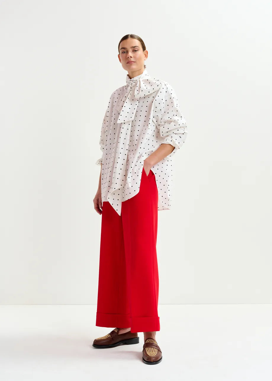 A person stands against a plain background in the Essentiel Antwerp Imoverit Polkadot Shirt - White & Black, bright red wide-leg trousers, and brown loafers, finished with a bow at the collar.