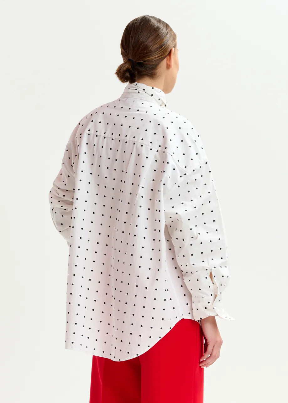 A person with brown hair in a low bun, facing away from the camera, wears the Essentiel Antwerp Imoverit Polkadot Shirt in white & black and bright red pants.