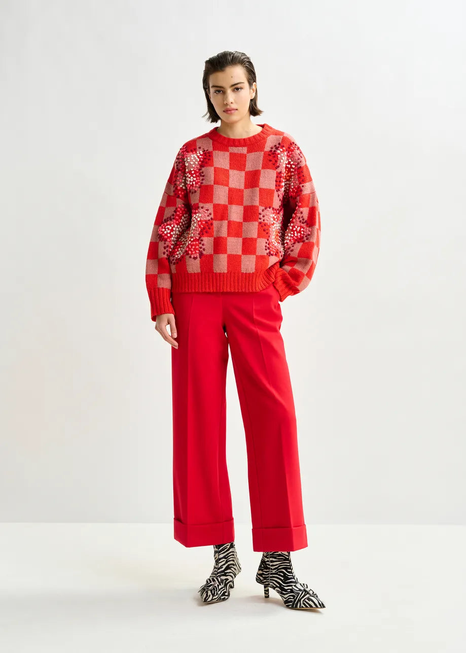A person stands against a plain background wearing Imo Cropped Trousers in red by Essentiel Antwerp, paired with a red checkered floral sweater and zebra-print ankle boots.