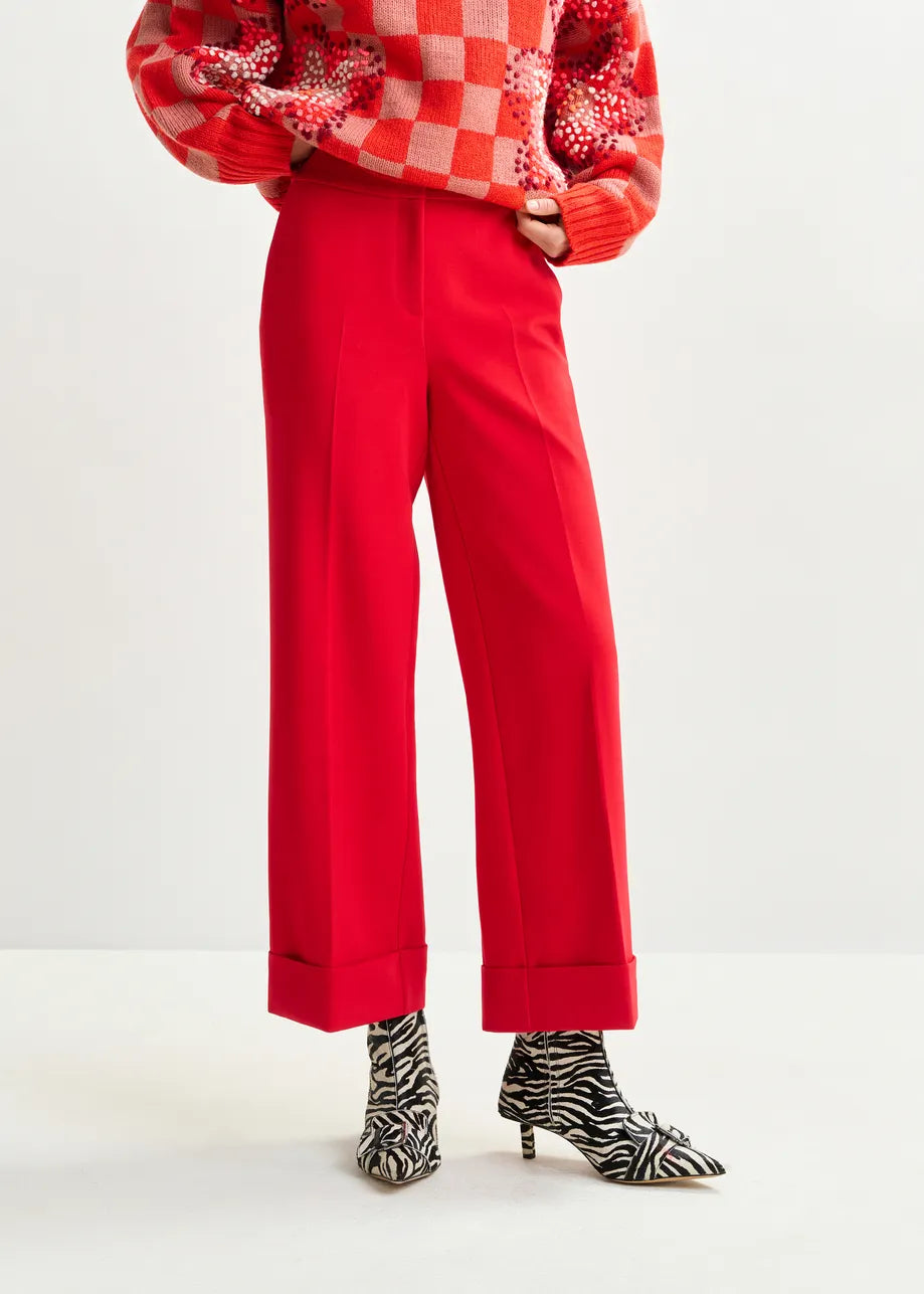A person wearing Essentiel Antwerp's Imo Cropped Trousers in red, paired with a red and pink checkered sweater and zebra print ankle boots, stands against a plain background.