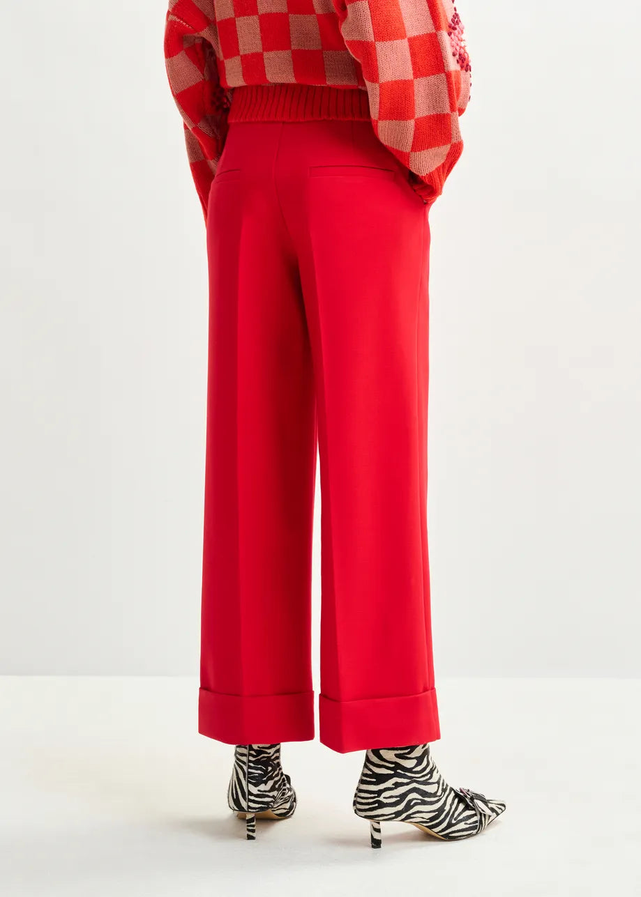 A person wearing Essentiel Antwerp’s Imo Cropped Trousers in red, paired with a red and pink checkered sweater and zebra-striped high-heeled ankle boots.