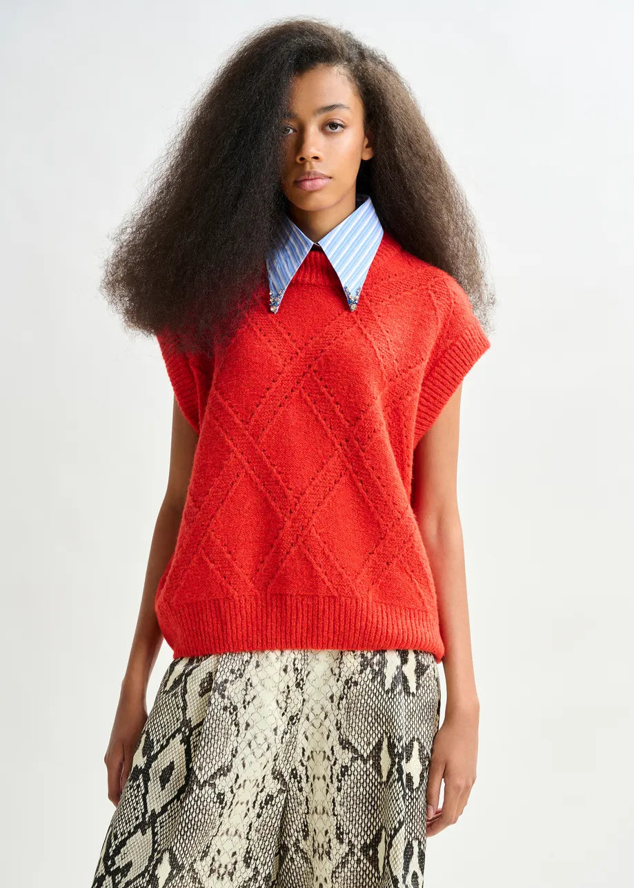 A person wearing the Essentiel Antwerp Import Cable-Knit Sweater in red over a blue collared shirt and a snakeskin-patterned skirt stands against a plain background.