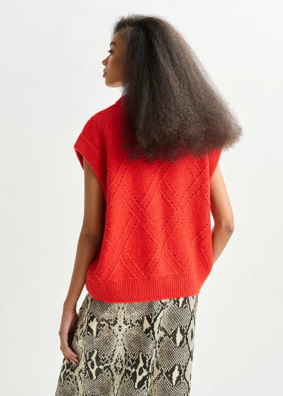 A woman stands facing away, wearing the Essentiel Antwerp Import Cable-Knit Sweater in red and a snakeskin-patterned skirt against a plain light background.
