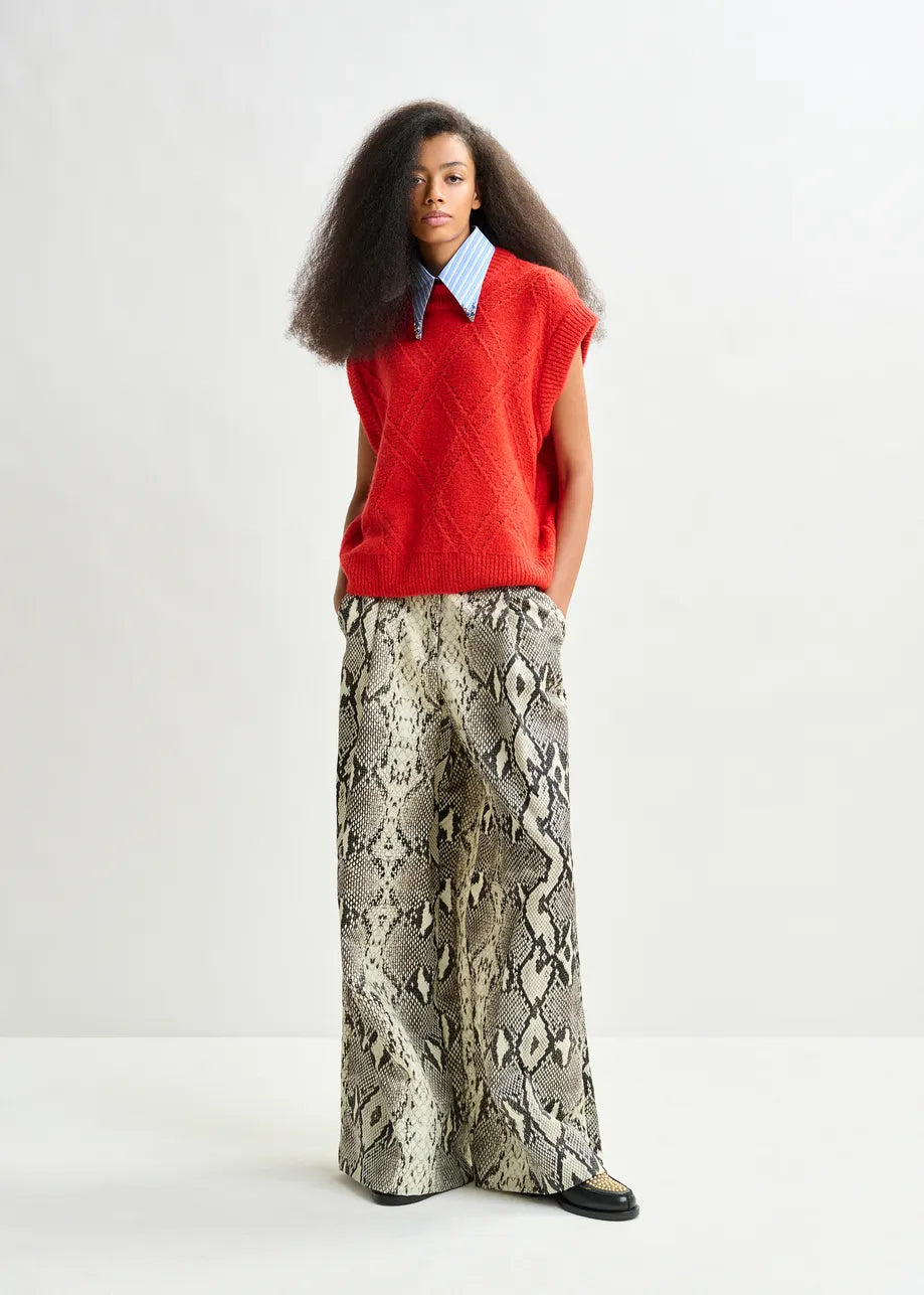 A person stands against a plain background wearing the Essentiel Antwerp Import Cable-Knit Sweater in red, paired with a blue collared shirt and wide-leg snakeskin-pattern pants.