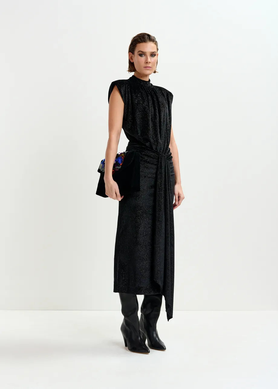 A woman stands against a plain white background wearing the Essentiel Antwerp Inche Midi Dress in black, paired with black heeled boots and a black clutch bag.