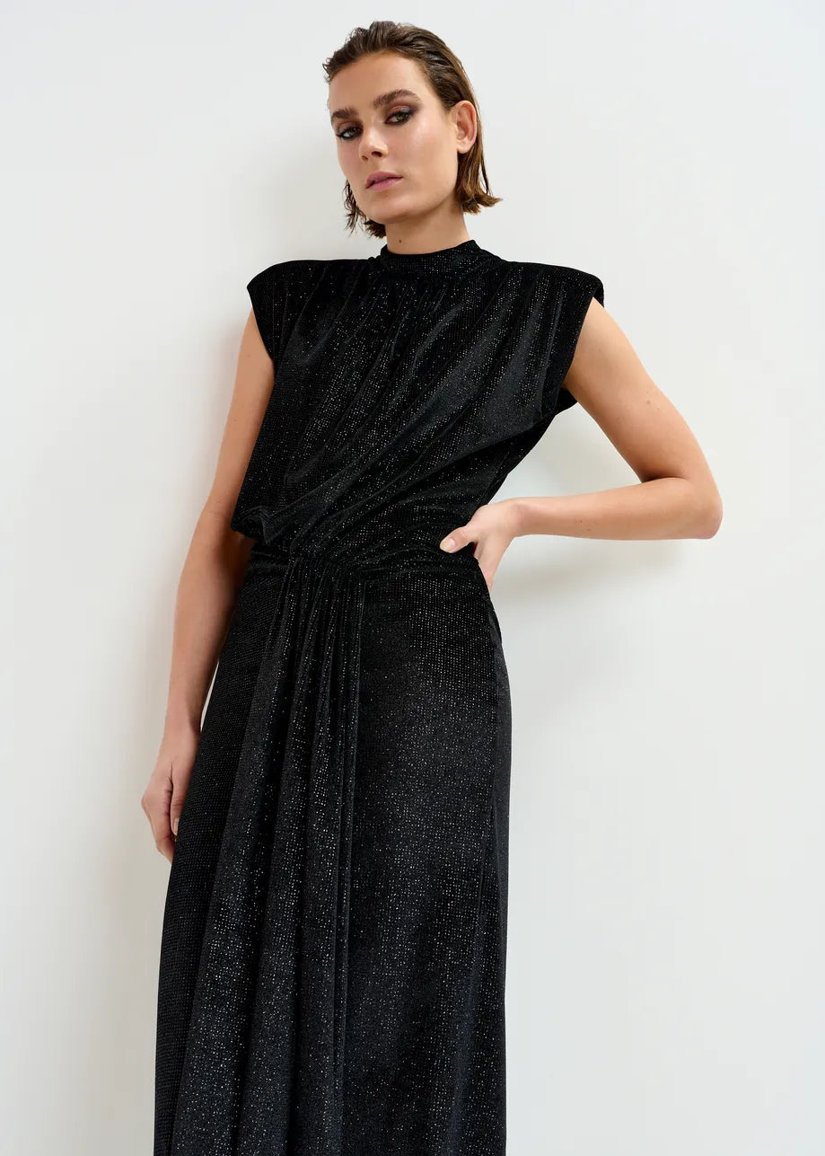 A person stands against a white wall wearing the Essentiel Antwerp Inche Midi Dress in black, featuring a sleeveless, high-neck design, shimmery stretch-jersey fabric, and draped waist detailing.