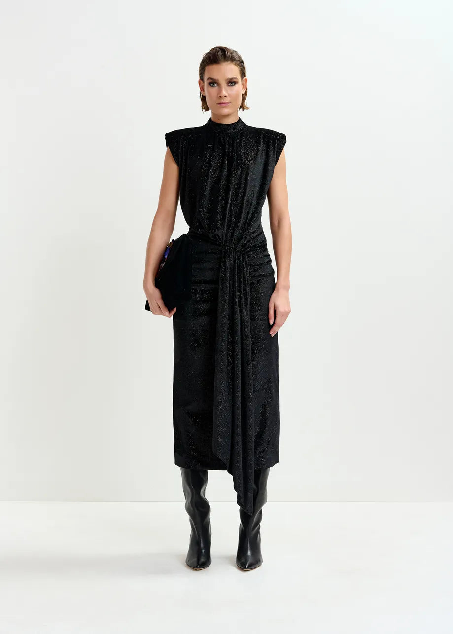 A person stands against a white background wearing the Essentiel Antwerp Inche Midi Dress in black, styled with knee-high black boots and a matching black clutch.