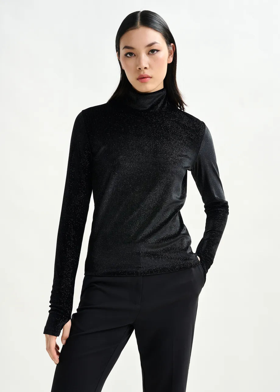 A woman poses against a plain background, wearing the Essentiel Antwerp Indienne Glitter Top in black with long sleeves and black stretch-jersey pants, one hand in her pocket.