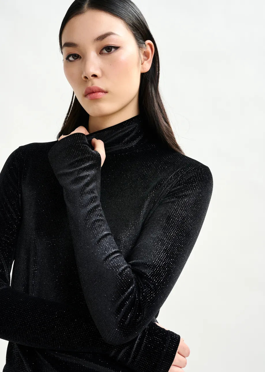 A woman with long dark hair wears the Indienne Glitter Top in black by Essentiel Antwerp, a fitted stretch-jersey turtleneck, standing against a plain light background and gazing at the camera with a neutral expression.