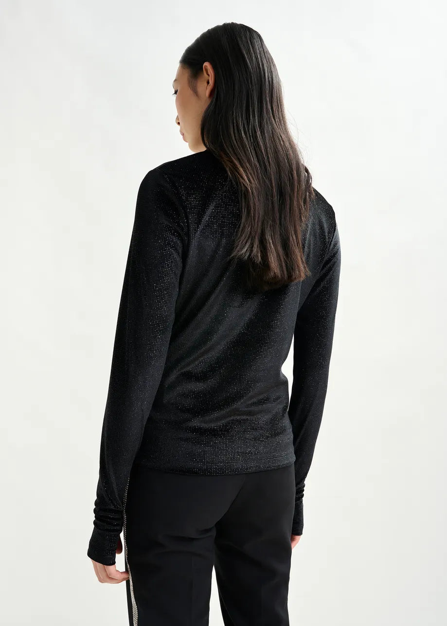 A person with long, dark hair faces away, wearing the Essentiel Antwerp Indienne Glitter Top in black stretch-jersey and black pants against a light background.