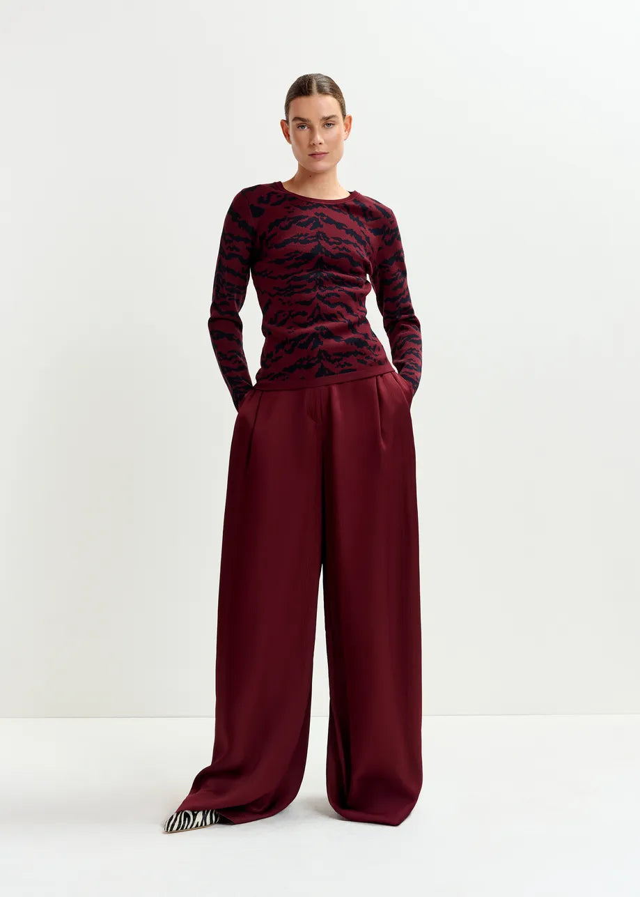 A woman stands against a plain background wearing the Inovember Satin Pants in burgundy by Essentiel Antwerp, styled with relaxed wide legs and her hands in her pockets.