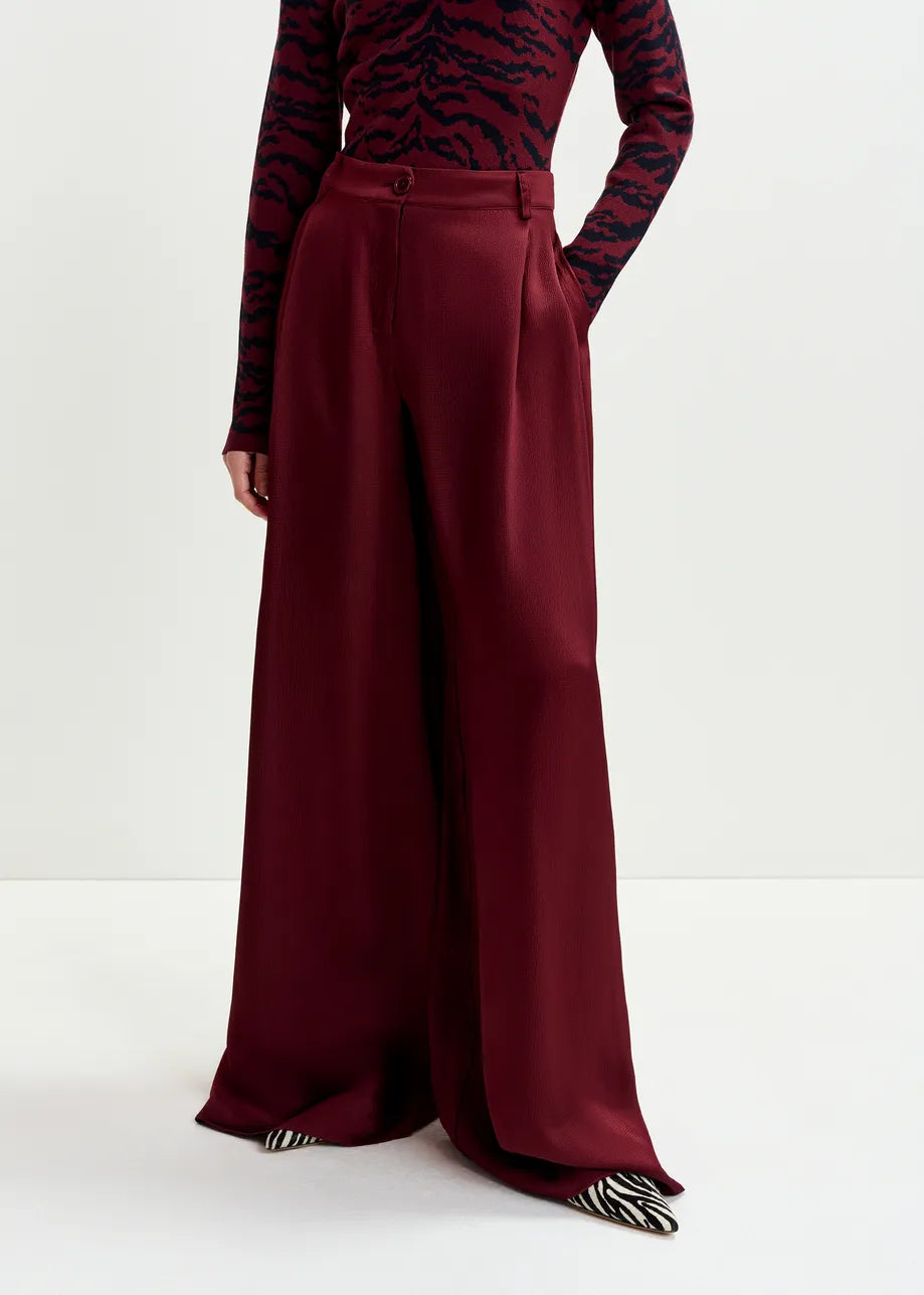 Model wears Inovember Satin Pants in burgundy by Essentiel Antwerp, paired with a long-sleeve dark red and black patterned top, both in flowing fabric, posed against a plain white background.