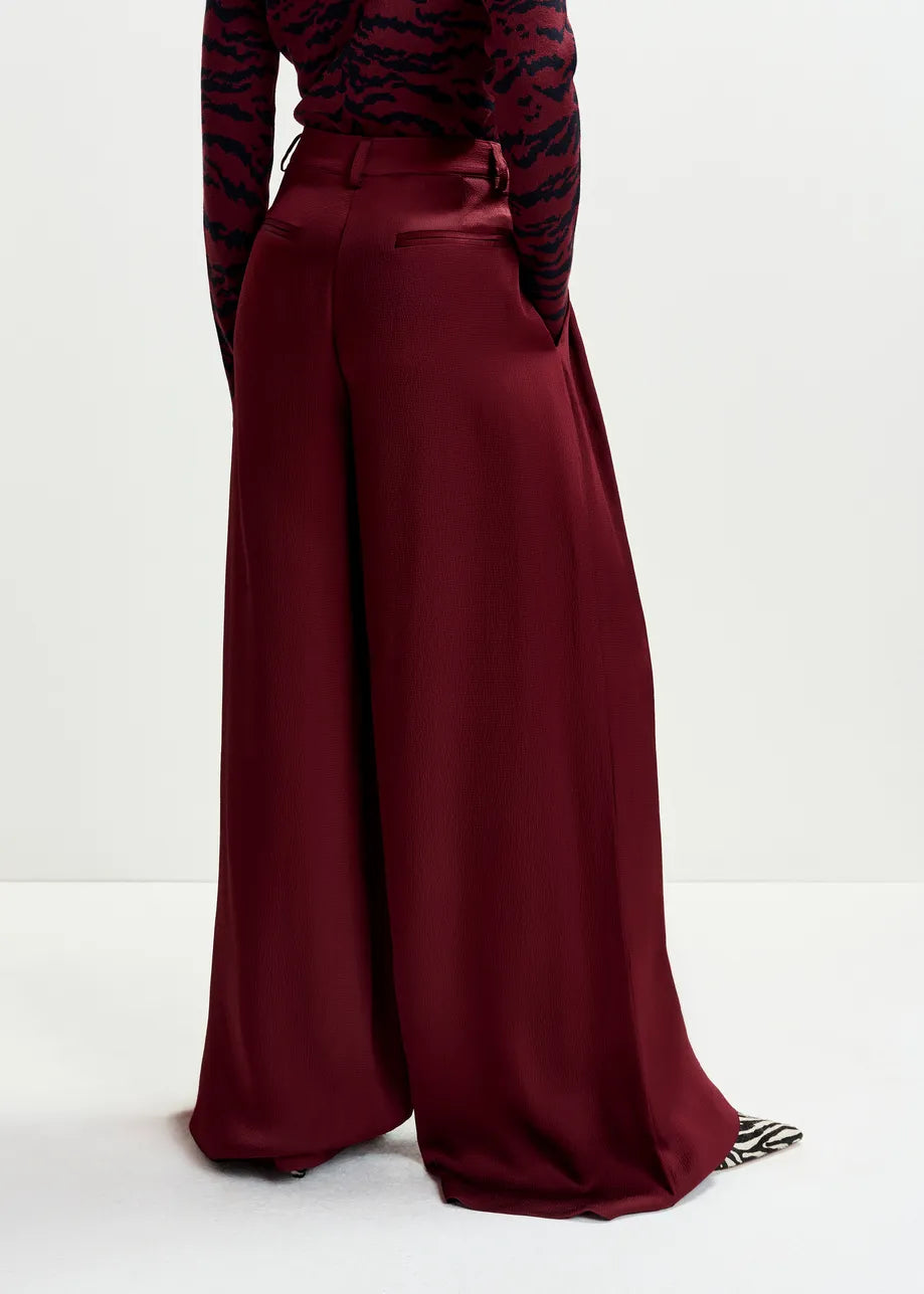 A person wears the Inovember Satin Pants in burgundy by Essentiel Antwerp—high-waisted, wide-leg trousers—in flowing fabric, paired with a long-sleeve patterned top and set against a plain light background.