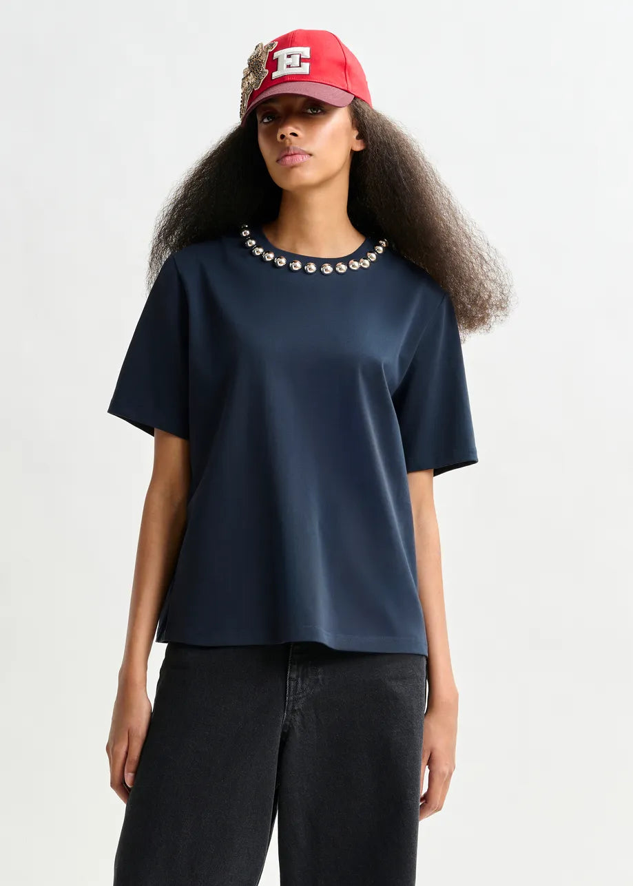 A person stands in front of a plain background wearing the Essentiel Antwerp Intenso Pearl-Embellished Tshirt in navy, paired with black jeans and a red baseball cap with an "E" emblem.