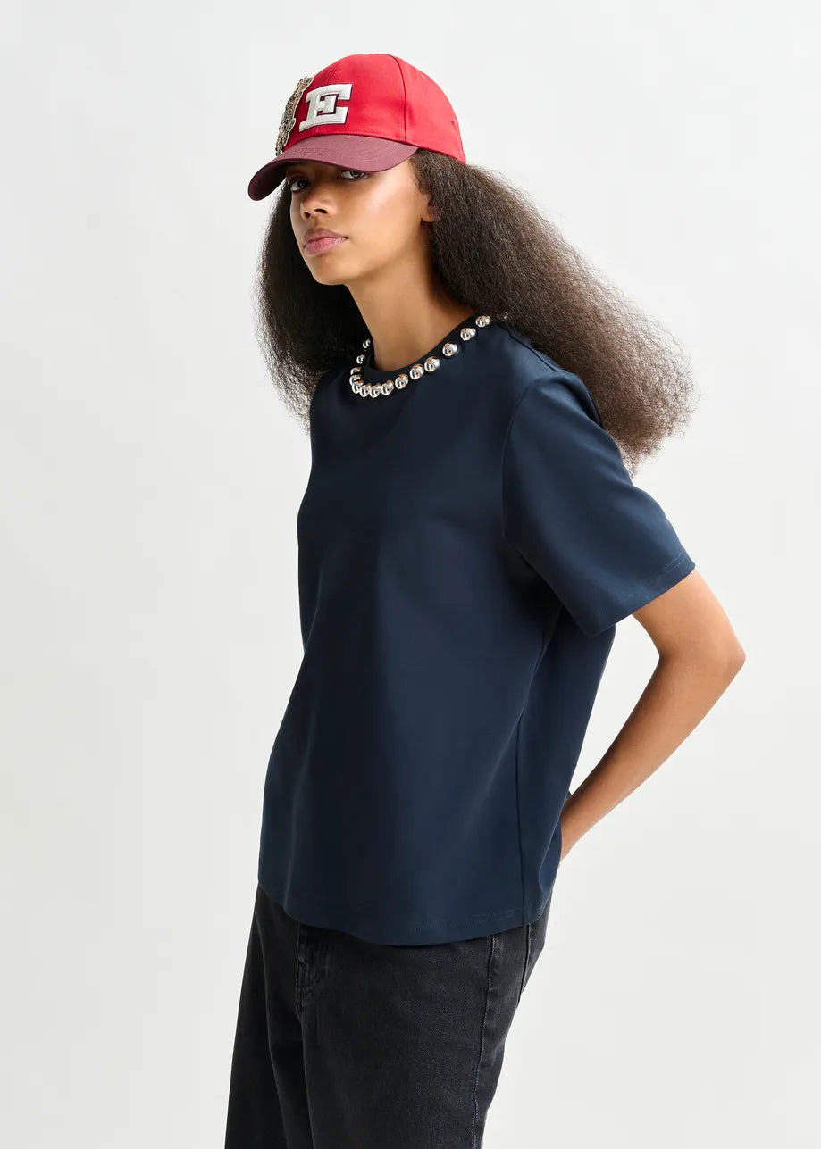 A person stands against a plain background wearing a red cap, black pants, and the Essentiel Antwerp Intenso Pearl-Embellished Tshirt in navy.