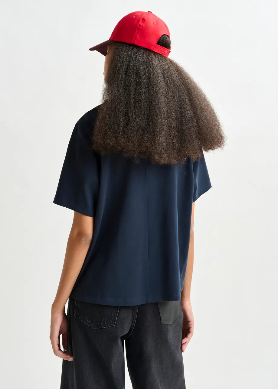 An individual stands with their back to the camera, wearing the Essentiel Antwerp Intenso Pearl-Embellished T-shirt in navy, paired with a red cap and black jeans, set against a plain background.
