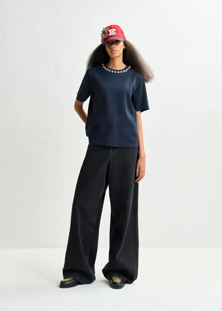 A person stands against a plain backdrop wearing a red cap with a flower accent, wide-leg black pants, black shoes, and the Essentiel Antwerp Intenso Pearl-Embellished Tshirt in navy with metallic pearls at the neckline.