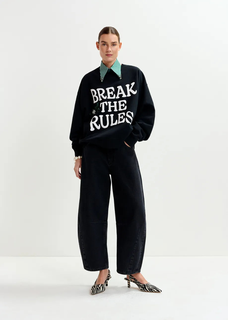 A person stands against a white background wearing a "BREAK THE RULES" sweatshirt, green collared shirt, Essentiel Antwerp’s Interstellar Barrel Leg Jeans in black, and zebra-print heels.