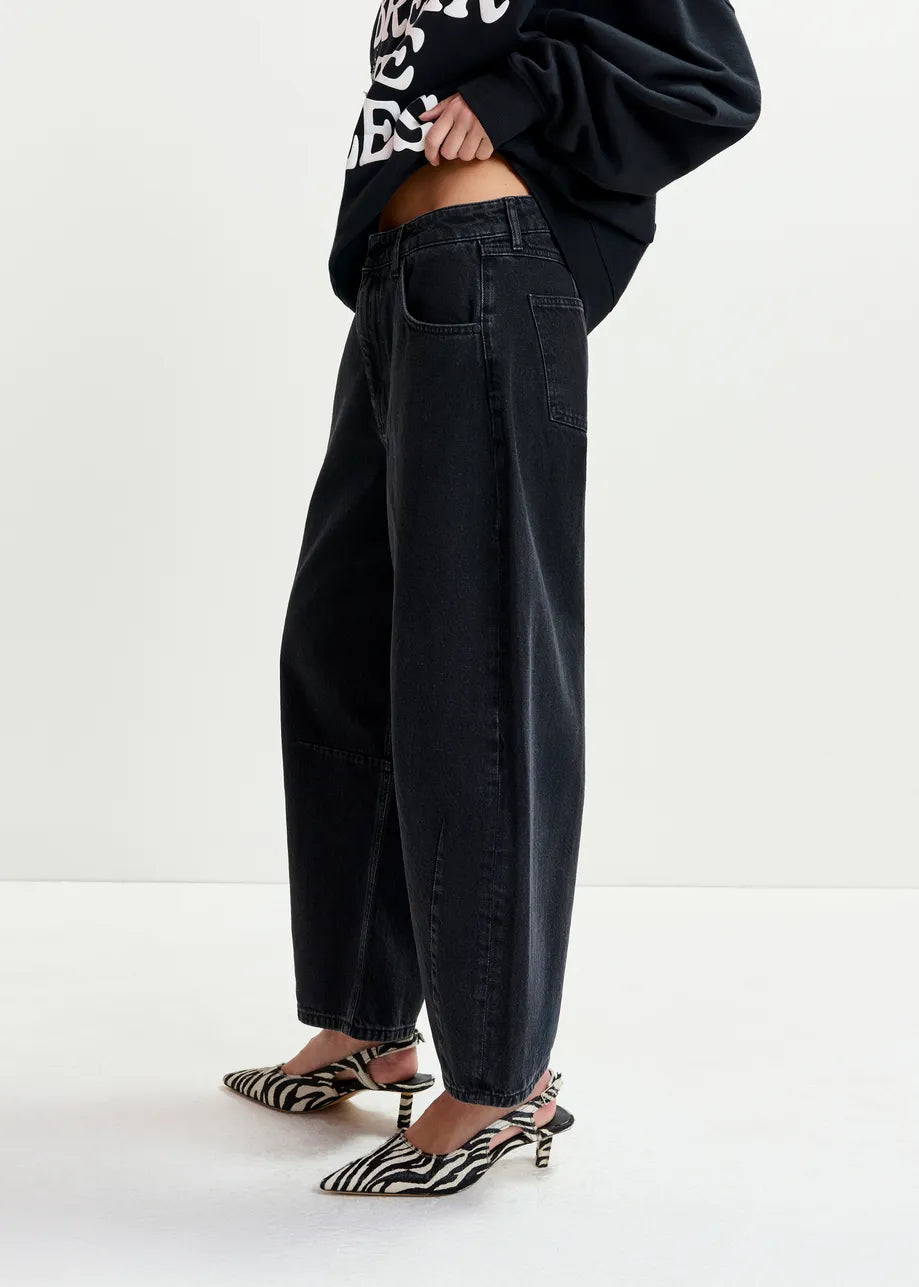 Person wearing Essentiel Antwerp Interstellar Barrel Leg Jeans in black, a black sweatshirt with white text, and zebra print heels, standing against a plain white background.