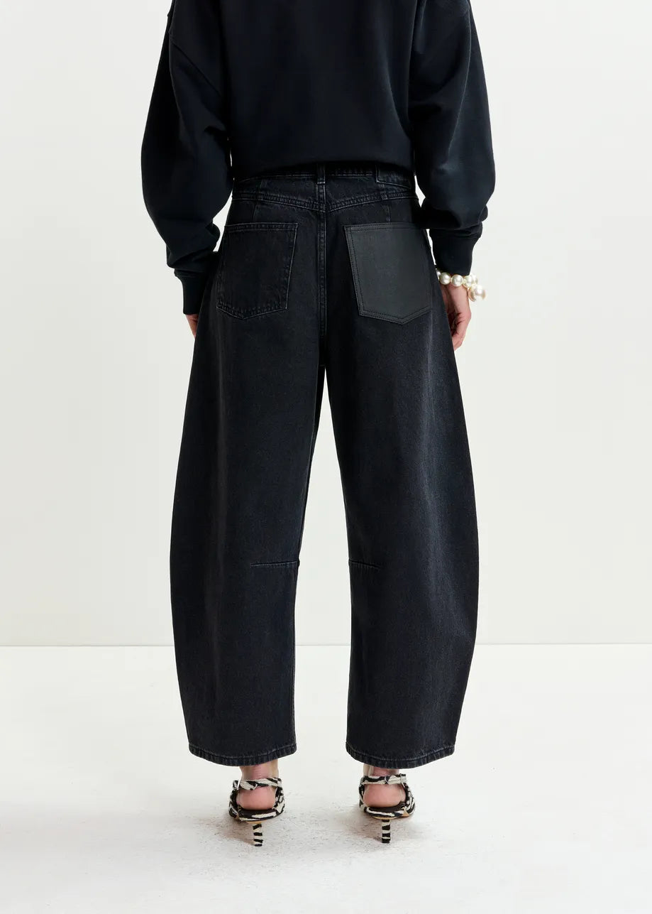 A person is seen from the back wearing Essentiel Antwerp's Interstellar Barrel Leg Jeans in black, featuring a contrasting black leather back pocket, styled with a black long-sleeve top and patterned open-toed heels.