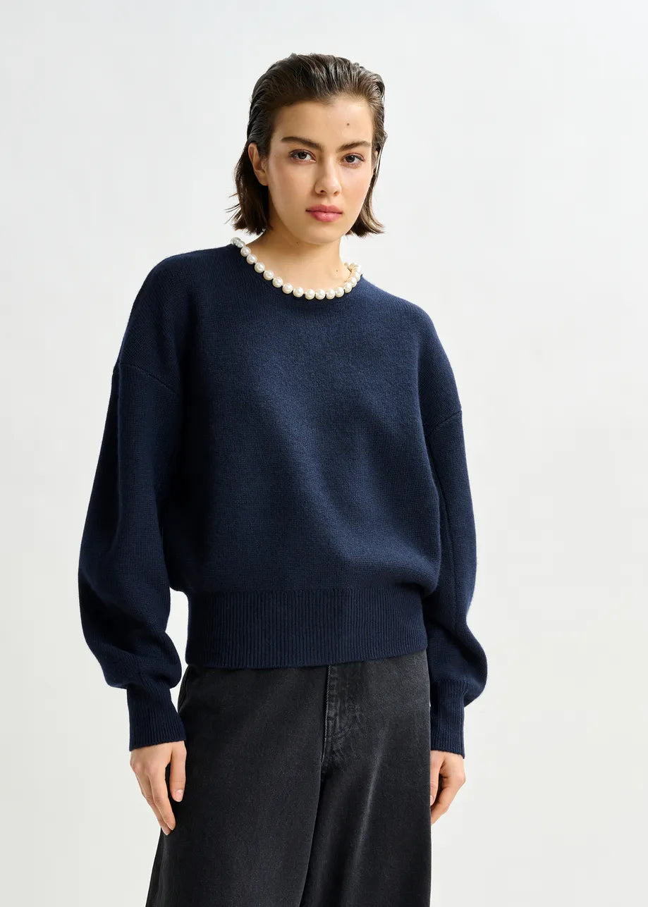 A person wears the Essentiel Antwerp Inti Pearl-Embellished Sweater in navy, styled with black pants, standing against a plain light background.