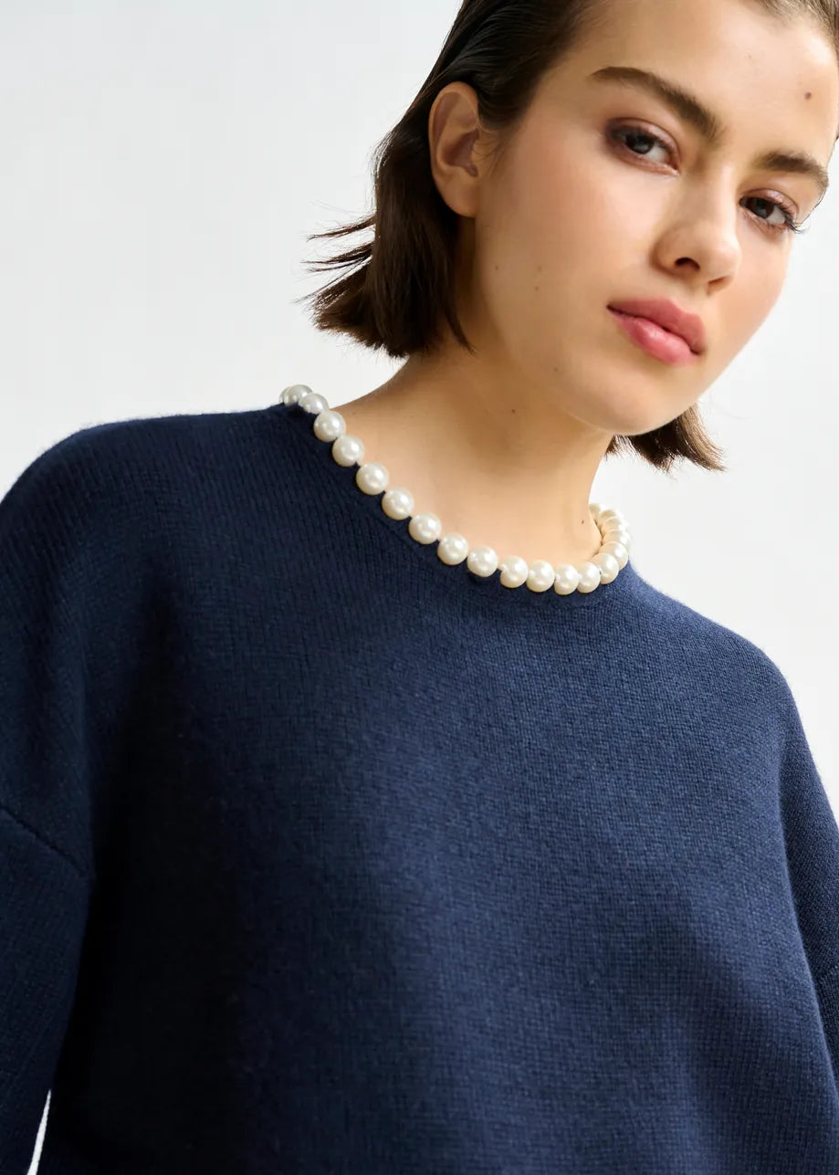 A person with short dark hair wears the Essentiel Antwerp Inti Pearl-Embellished Sweater in navy, paired with a pearl necklace, looking slightly to the side against a plain background.