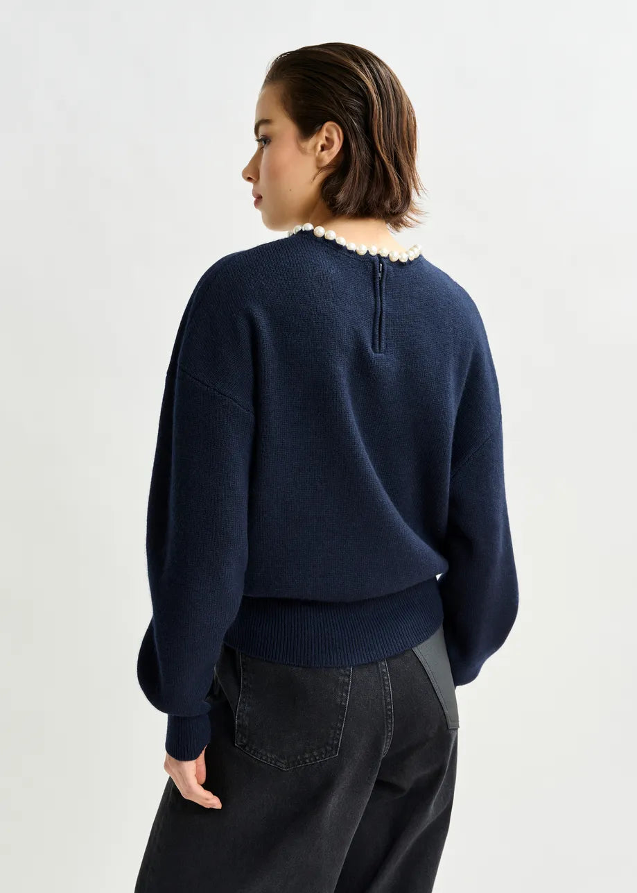 A person faces away, wearing the Essentiel Antwerp Inti Pearl-Embellished Sweater in navy and black jeans, highlighting its relaxed fit and pearl-detailed neckline against a plain light background.