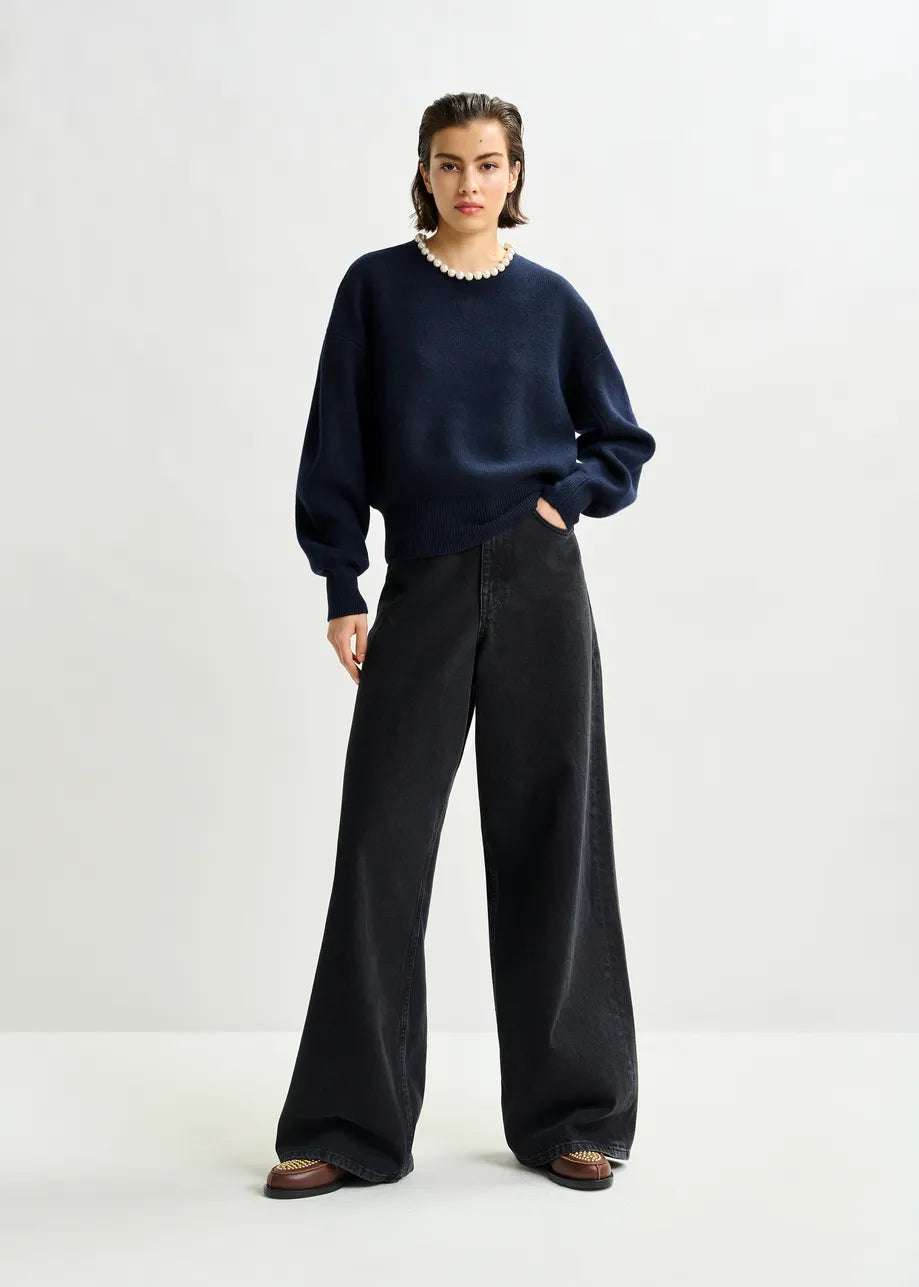 A person stands against a plain background wearing the Essentiel Antwerp Inti Pearl-Embellished Sweater in navy, paired with black wide-leg pants, brown shoes, and a pearl necklace.