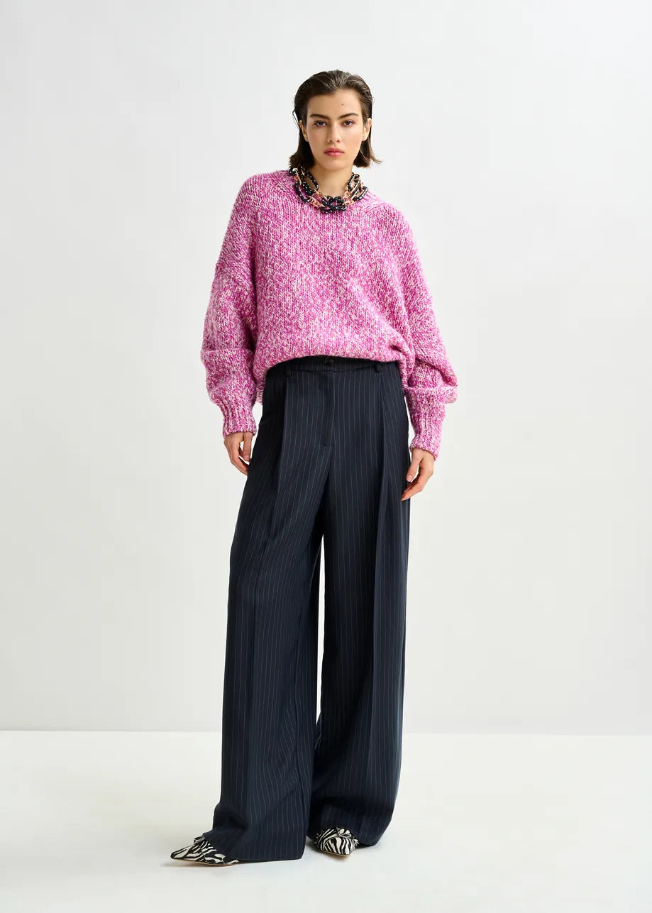 A person stands against a plain background wearing a loose pink knit sweater, pointed black-and-white patterned shoes, and Essentiel Antwerp's Invest Wide-Leg Trousers in navy pinstripe with a high waist.