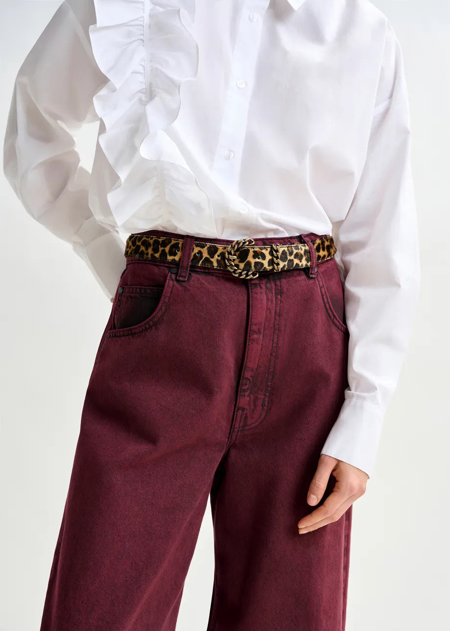 A person wearing a white ruffled shirt tucked into high-waisted maroon jeans, styled with the Essentiel Antwerp Ipony Belt in leopard print, featuring a sculptural metal buckle.
