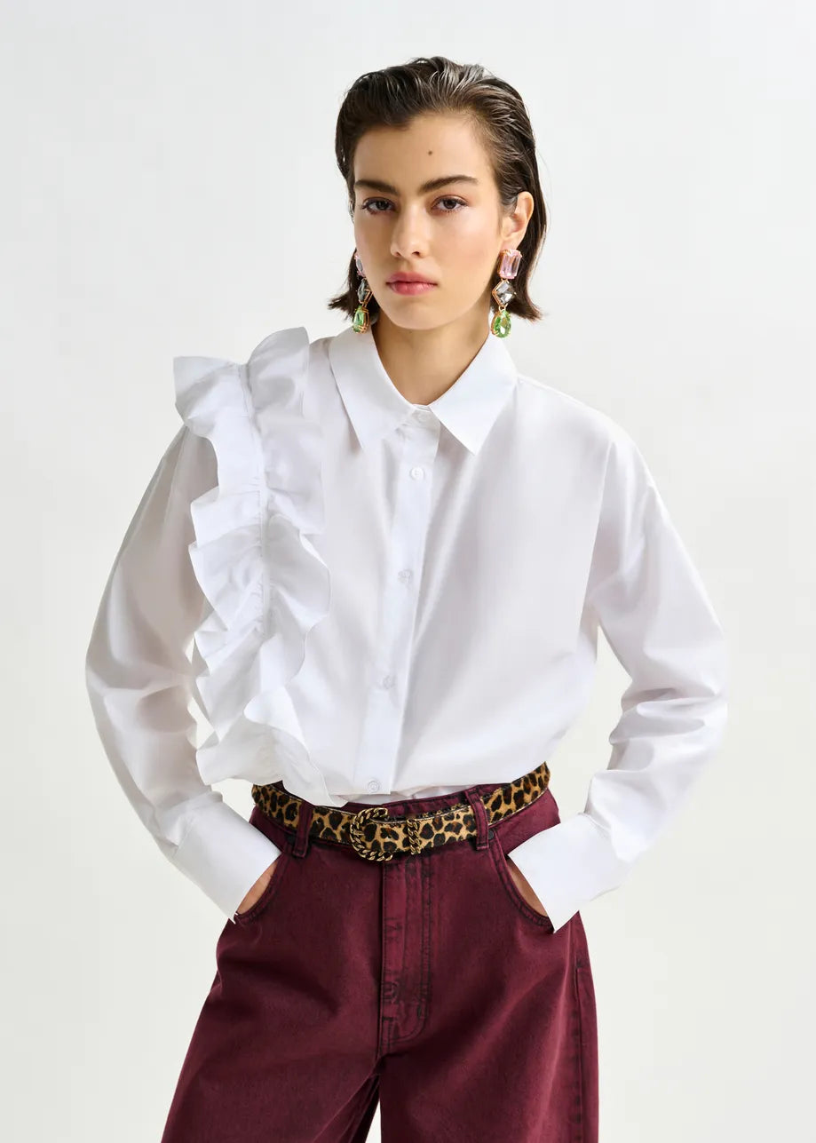 A person wears a white ruffled shoulder shirt, maroon high-waisted pants, statement earrings, and the Essentiel Antwerp Ipony Belt - Leopard with a sculptural metal buckle, standing against a plain background.
