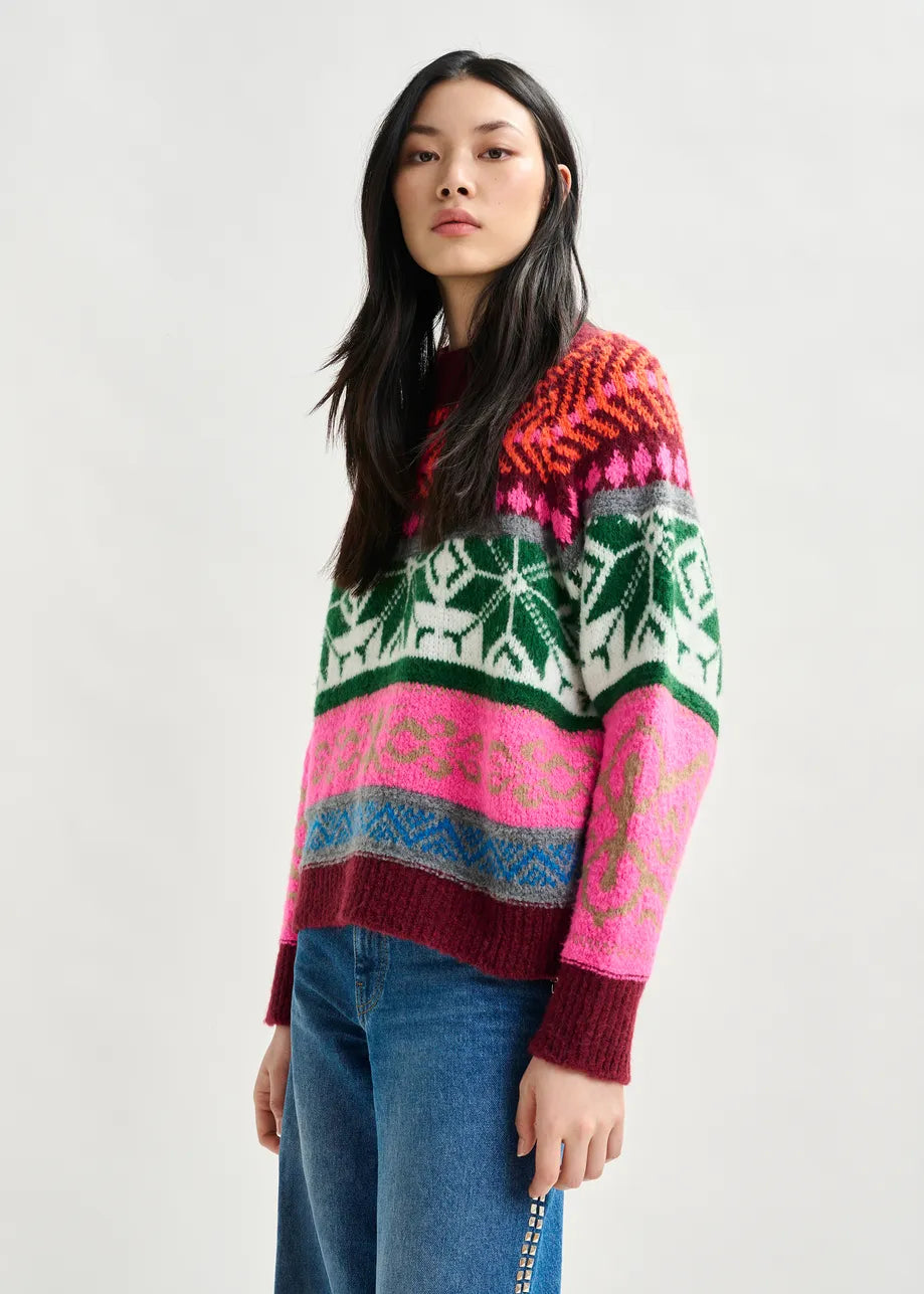 A person wears the Essentiel Antwerp Irelando Jacquared Knit - Burgundy sweater, featuring red, pink, green, and blue designs, paired with blue jeans, standing against a plain light background.