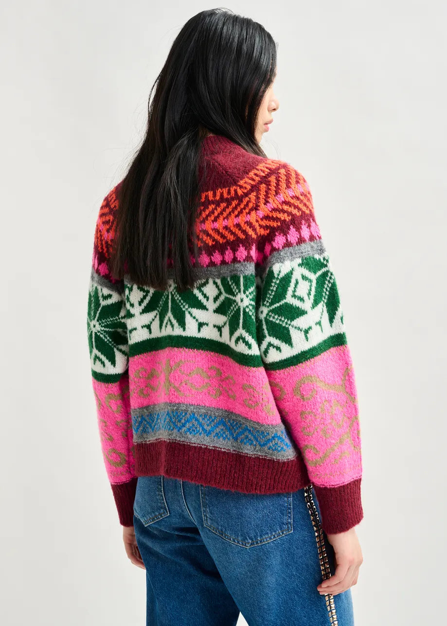 A person stands with their back to the camera, wearing the Essentiel Antwerp Irelando Jacquared Knit in Burgundy featuring vibrant pink, green, red, and white jacquard patterns, paired with blue jeans against a light background.