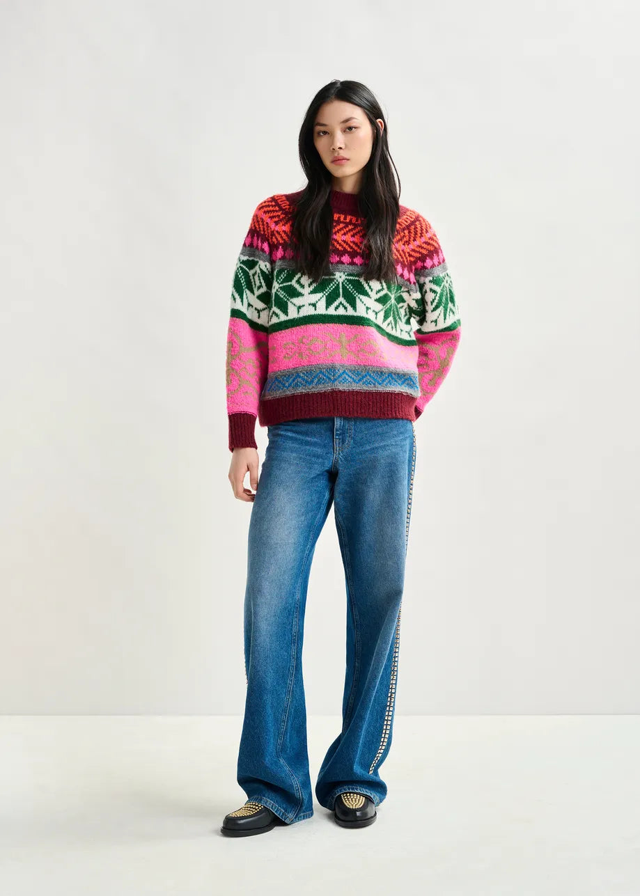 A woman stands against a plain background wearing the Essentiel Antwerp Irelando Jacquared Knit in burgundy, paired with blue wide-leg jeans and black-and-white shoes.