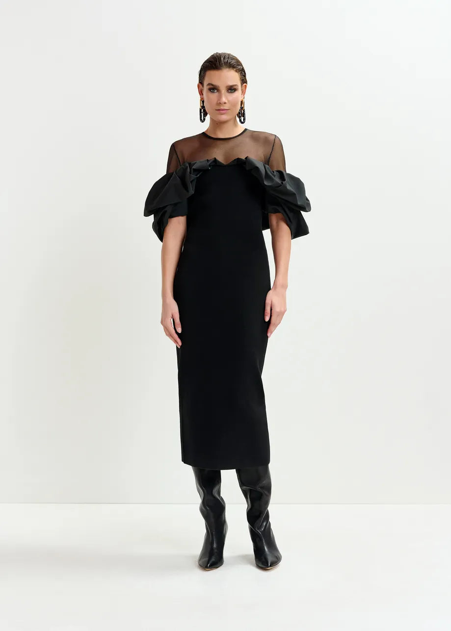 A woman models the Essentiel Antwerp Isatis Ruffle Dress in black, featuring off-shoulder ruffle sleeves and a sheer neckline, paired with statement earrings and knee-high boots—ideal for chic evening wear against a white backdrop.
