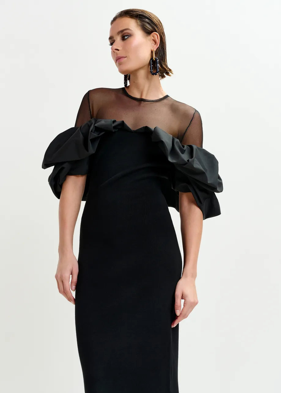 A woman models the Essentiel Antwerp Isatis Ruffle Dress in black, featuring sheer mesh, ruffle shoulders, and dramatic sleeves—an elegant evening choice—against a white background.