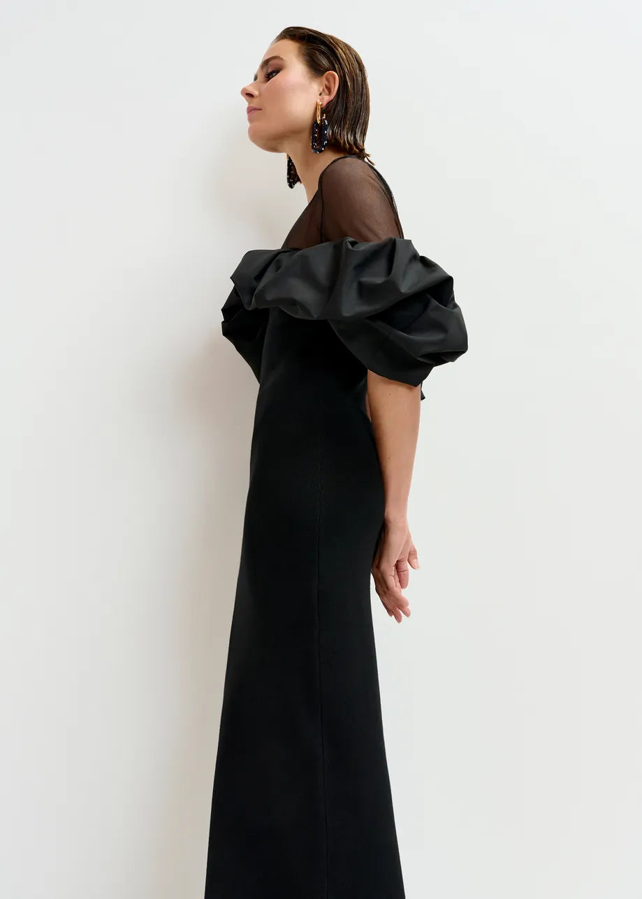 A woman in the Isatis Ruffle Dress - Black by Essentiel Antwerp—a black off-the-shoulder gown with sheer puff sleeves—stands in profile against a plain white background.