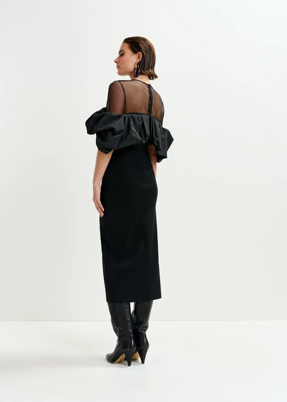 A woman faces away in the Essentiel Antwerp Isatis Ruffle Dress - Black, featuring ruffled sleeves and a sheer top, styled with black heeled boots against a plain white background—an elegant evening look.